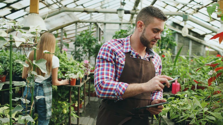 Inside a well-lit greenhouse with a transparent roof, two individuals are engaged in garden maintenance and planning activities. In the foreground, a woman with dark braided hair and glasses, wearing